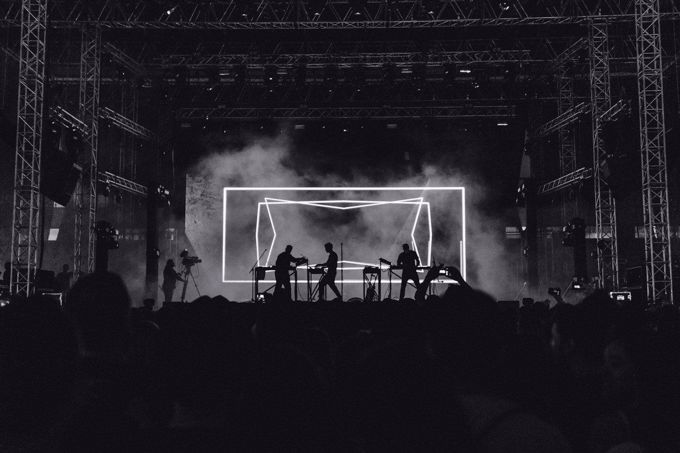 A band performing inside a frame of white light on a festival stage, haze rising.